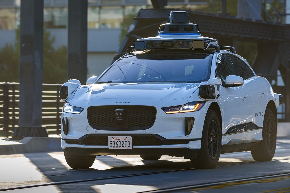 A white Waymo Jaguar I-Pace with roof-mounted sensors driving on a city street