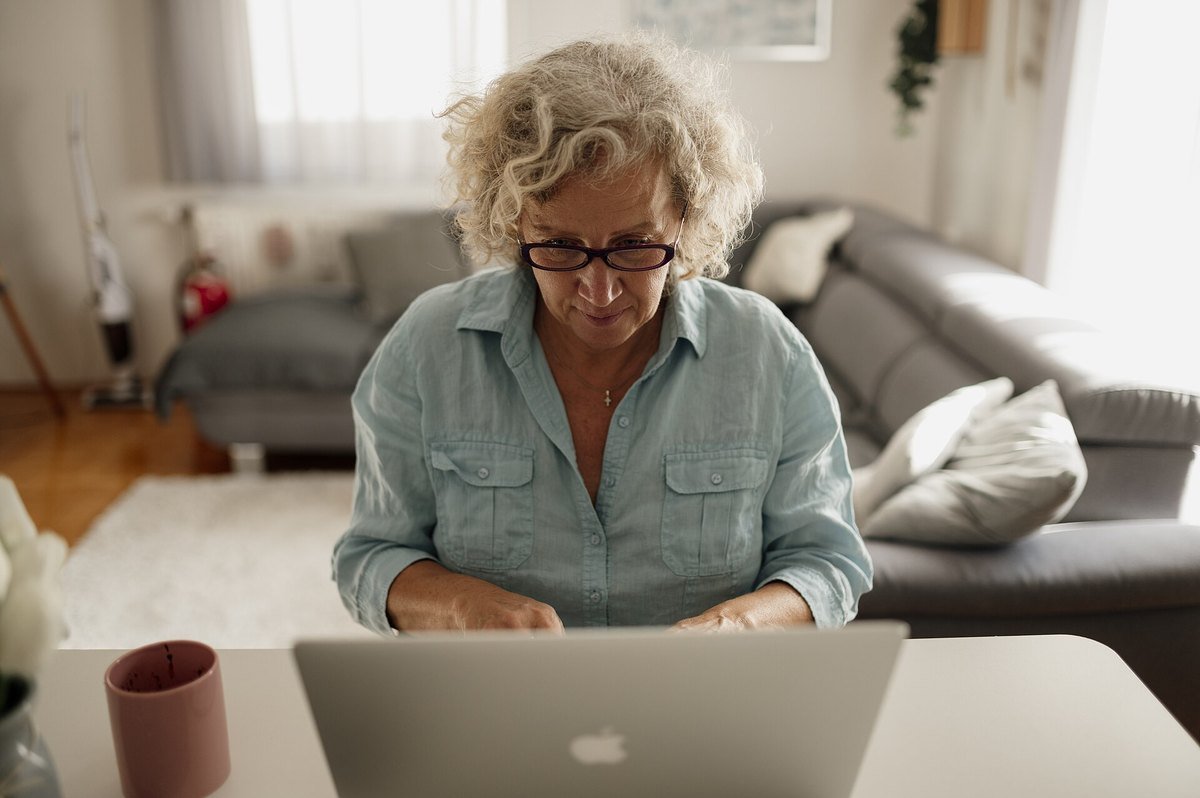 A woman in glasses leaning toward her laptop screen in a bright living room, reading carefully.