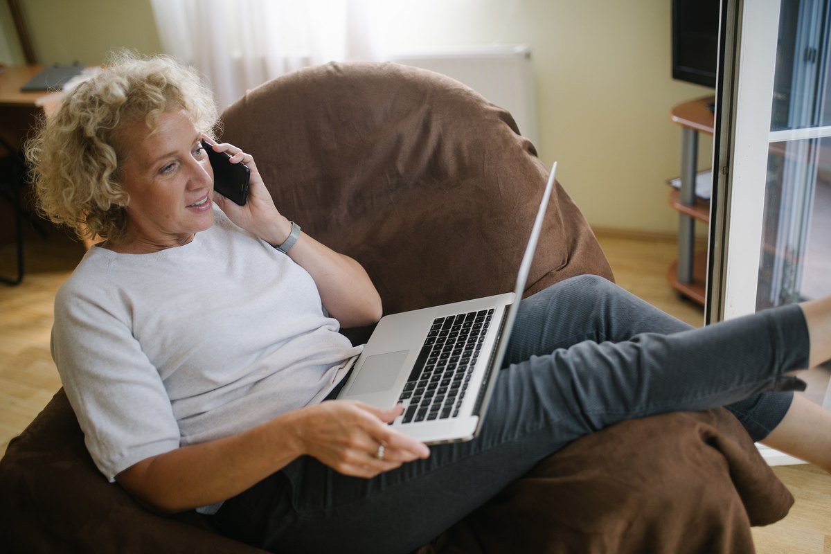 A woman in an armchair talking on a phone with a laptop open on her lap.