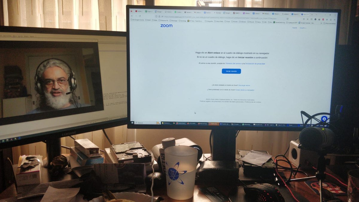 A desk with two screens: the Zoom sign-in page on one, and a man in headphones on a Zoom call on the other