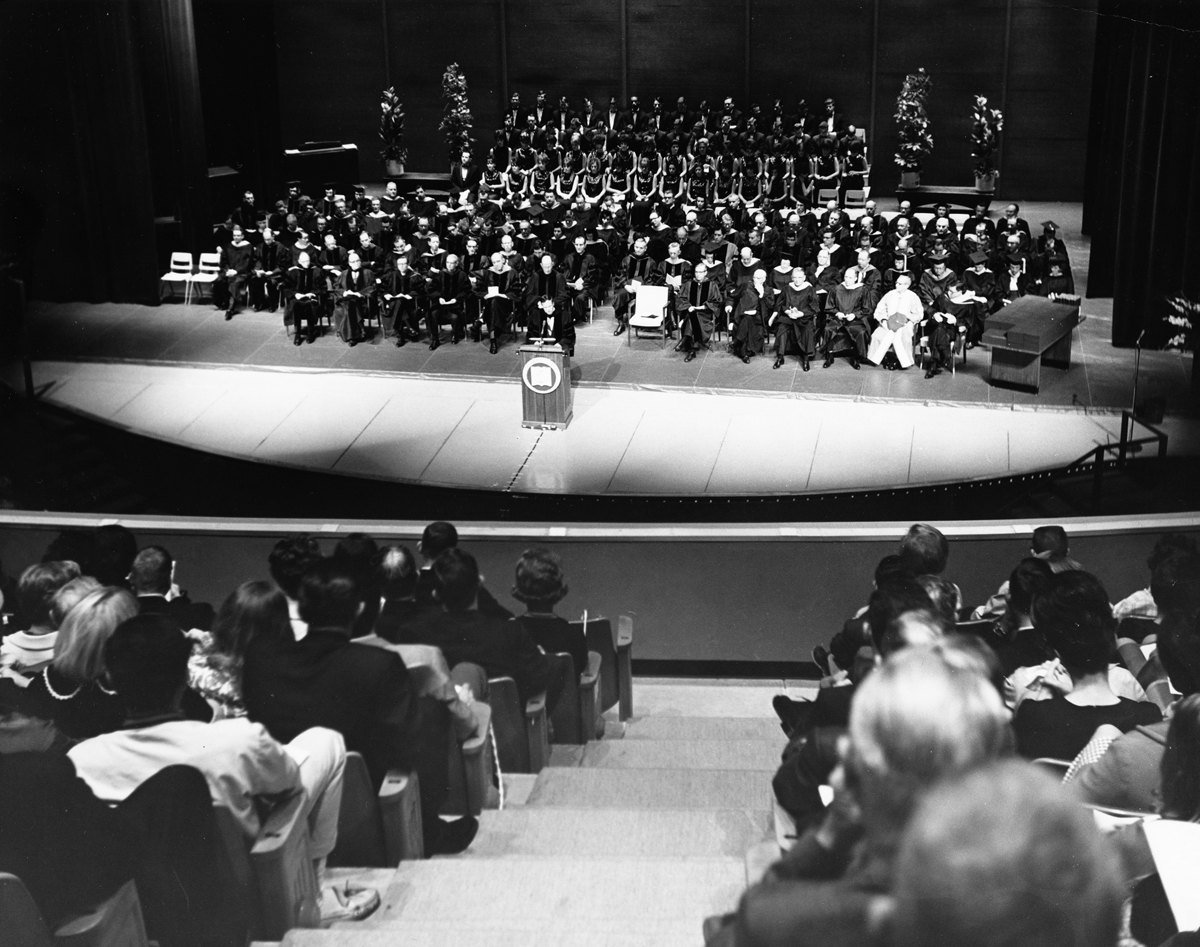 Black and white photo of a 1950s Arlington State College graduation ceremony, a stage with faculty and rows of graduates seen from the back of the hall