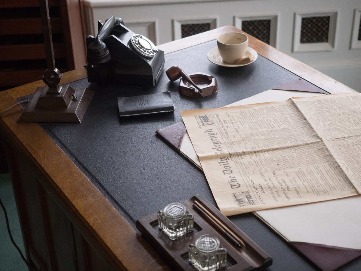 A wooden office desk with a black rotary telephone, a desk lamp, a cup and saucer, glass inkwells and an open newspaper on a leather blotter