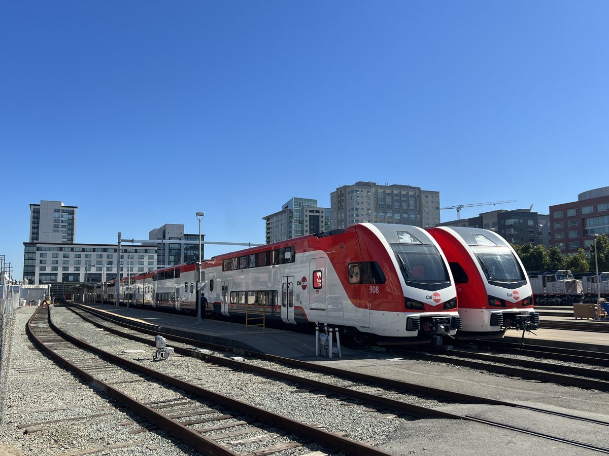 Two red and white Caltrain electric trains parked at the San Francisco 4th and King station under a clear blue sky, with apartment and office buildings behind