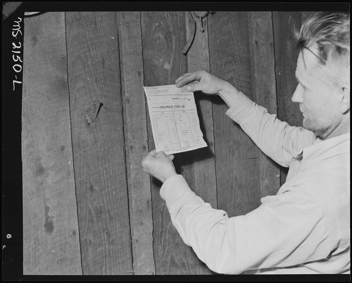 Black and white 1946 photo of coal miner Charlie Davis holding his Coleman Fuel Company pay slip flat against a wooden wall.