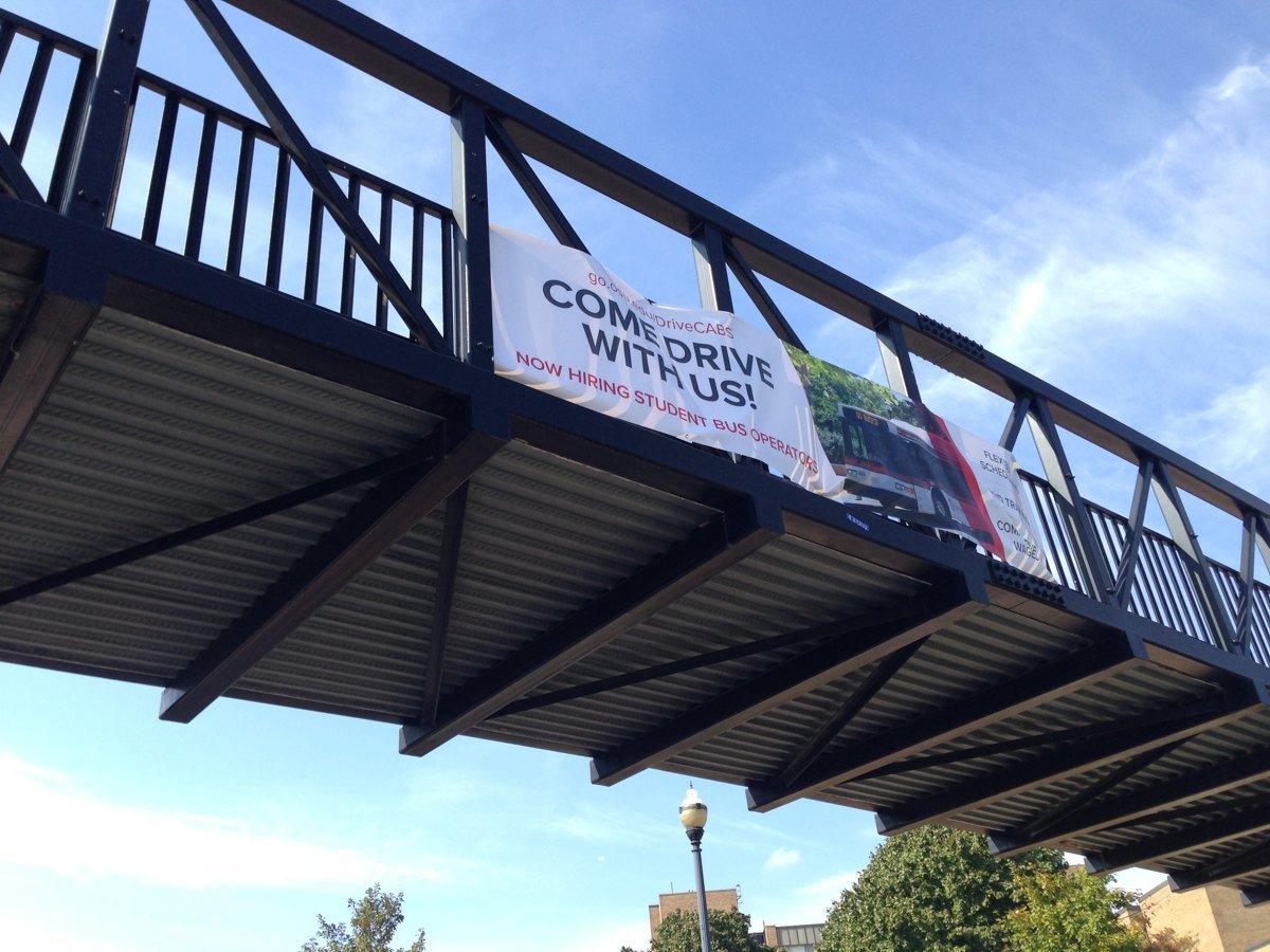 A pedestrian bridge seen from below against a blue sky, with a white banner hanging on its railing that reads Come drive with us.