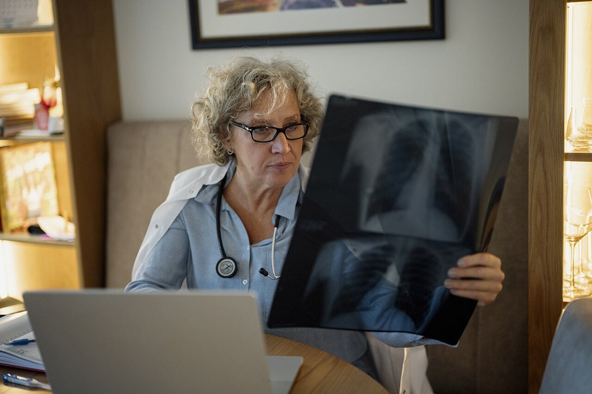 A woman with glasses and a stethoscope holds up a chest X-ray while sitting at a home desk with a laptop open in front of her.