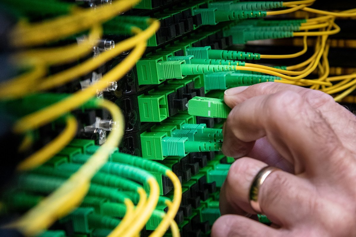 Close-up of a technician's hand plugging a green fiber-optic connector into a dense patch panel threaded with yellow fiber cables.