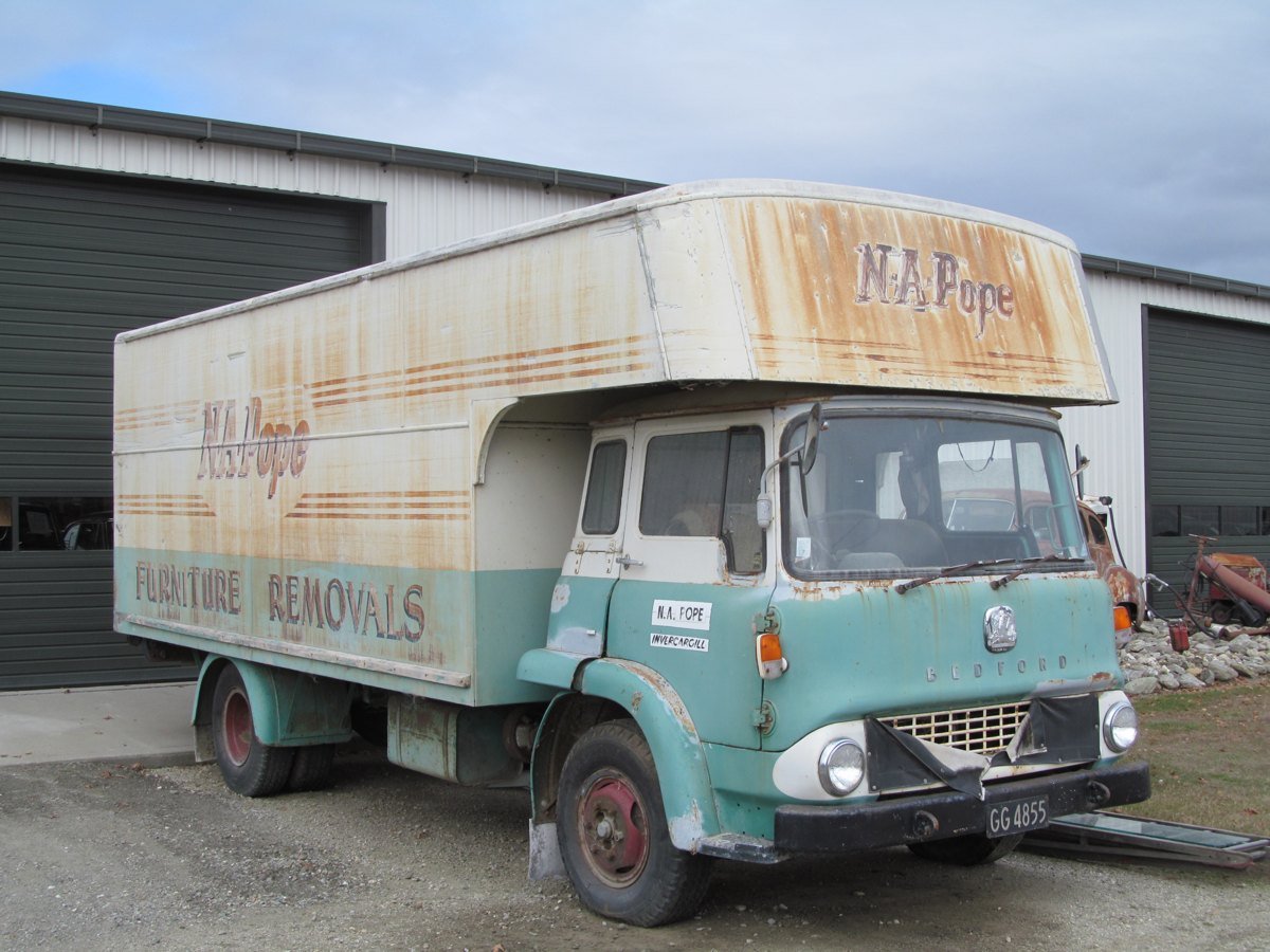 A faded teal and cream Bedford furniture removals truck with rusted paintwork parked outside a metal shed