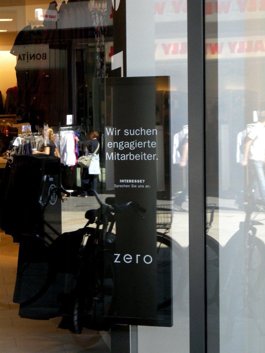 Black panel in a clothing shop window reading, in German, Wir suchen engagierte Mitarbeiter, Interesse? Sprechen Sie uns an, with the street and bicycles reflected in the glass