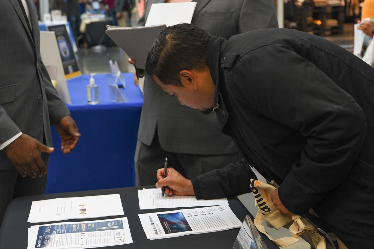 A job seeker bends over a table at a 2022 job fair, writing on a paper sign-in sheet next to employer handouts