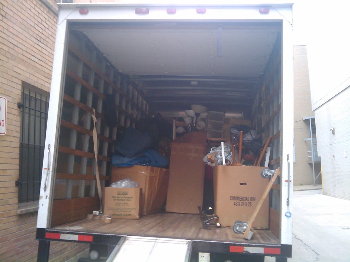 The open back of a rental moving truck parked beside a brick building, loaded with cardboard boxes, a wardrobe box, lamps and furniture under blue moving blankets