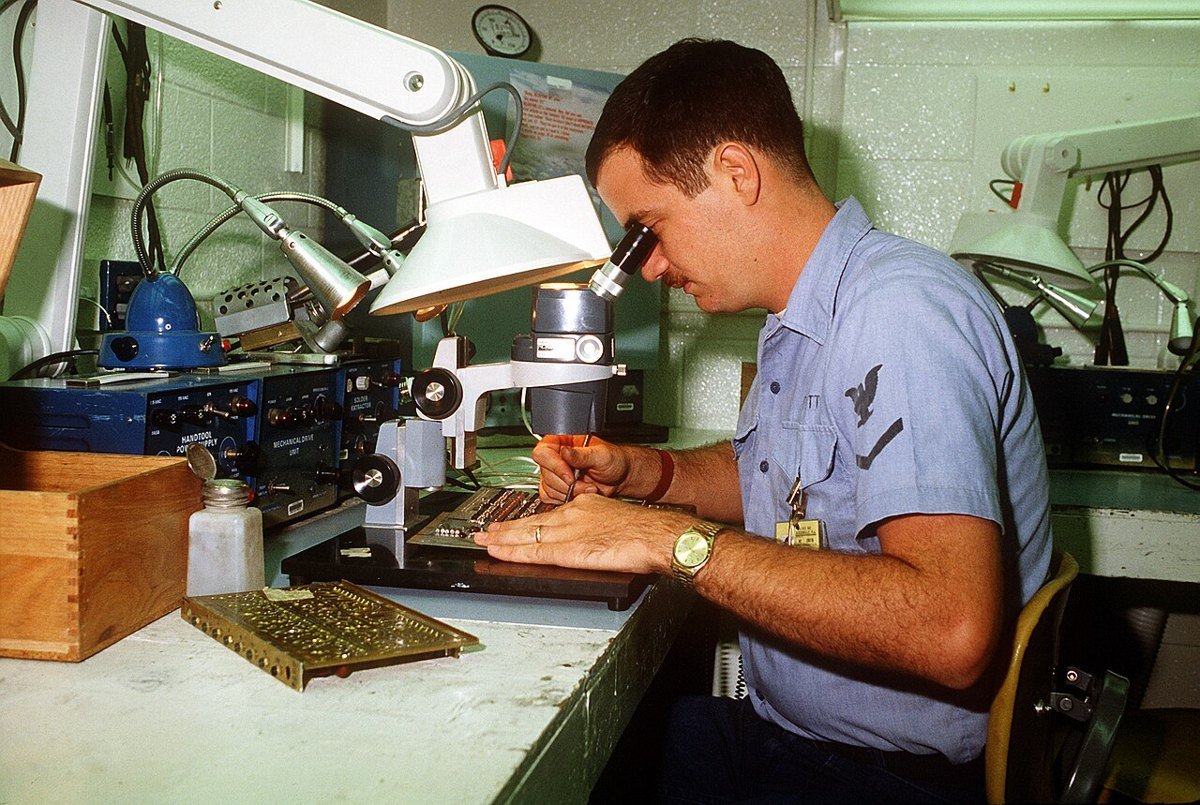 A Navy aviation electronics technician looks through a bench magnifier while working on a printed circuit board in a repair shop.