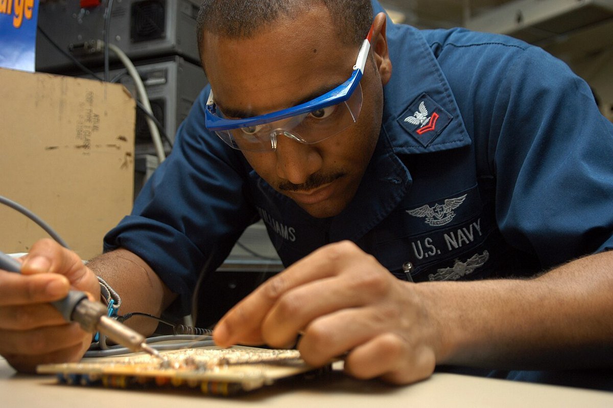 A Navy aviation electronics technician wearing safety glasses solders a component on a circuit card at a workbench.