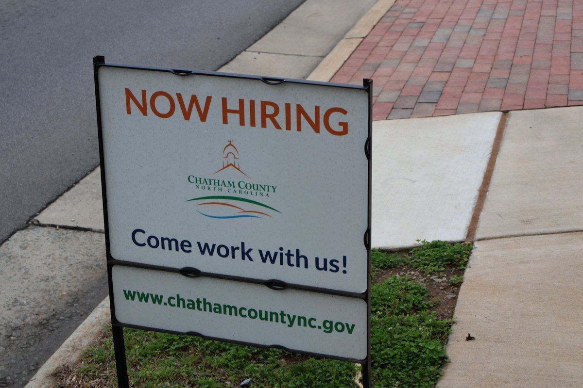 White roadside sign reading Now Hiring, Chatham County, Come work with us, with the county website address, planted in grass beside a sidewalk