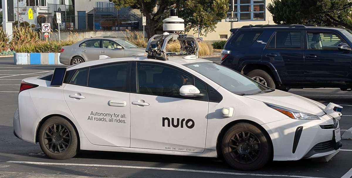 A white Nuro autonomous test car, a modified Toyota Prius with a roof sensor rack, parked in a San Francisco lot.