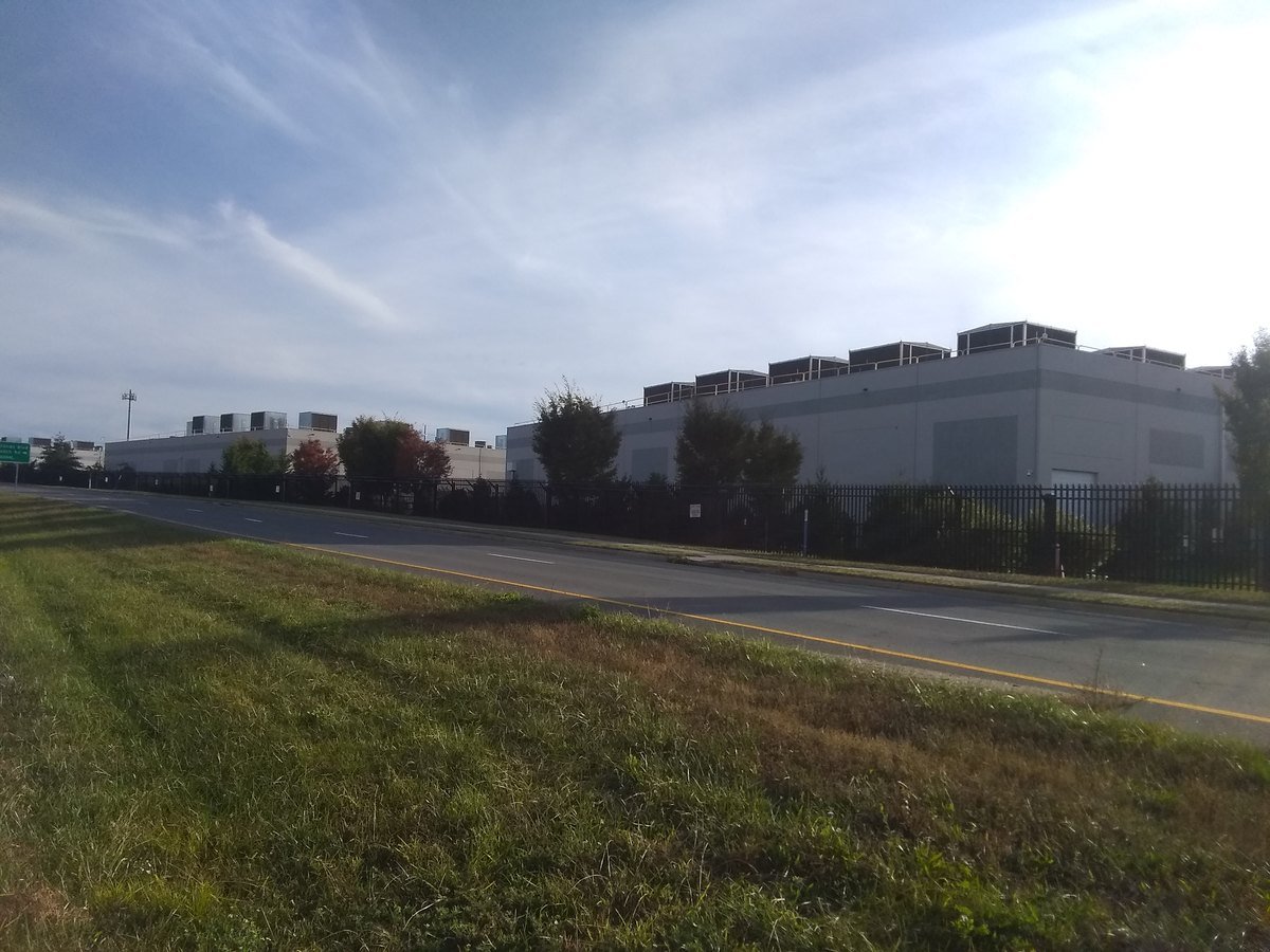 A long, windowless Amazon Web Services data center building behind a fence and a grass field along a road in Ashburn, Virginia.