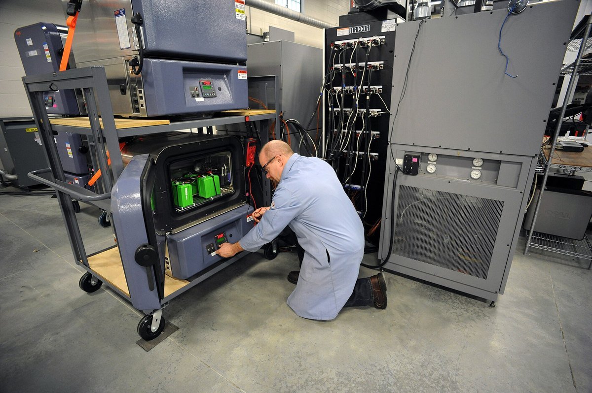 A technician in a gray lab coat kneels to adjust controls on a battery test chamber holding green battery modules, next to tall racks of test cables in a laboratory.
