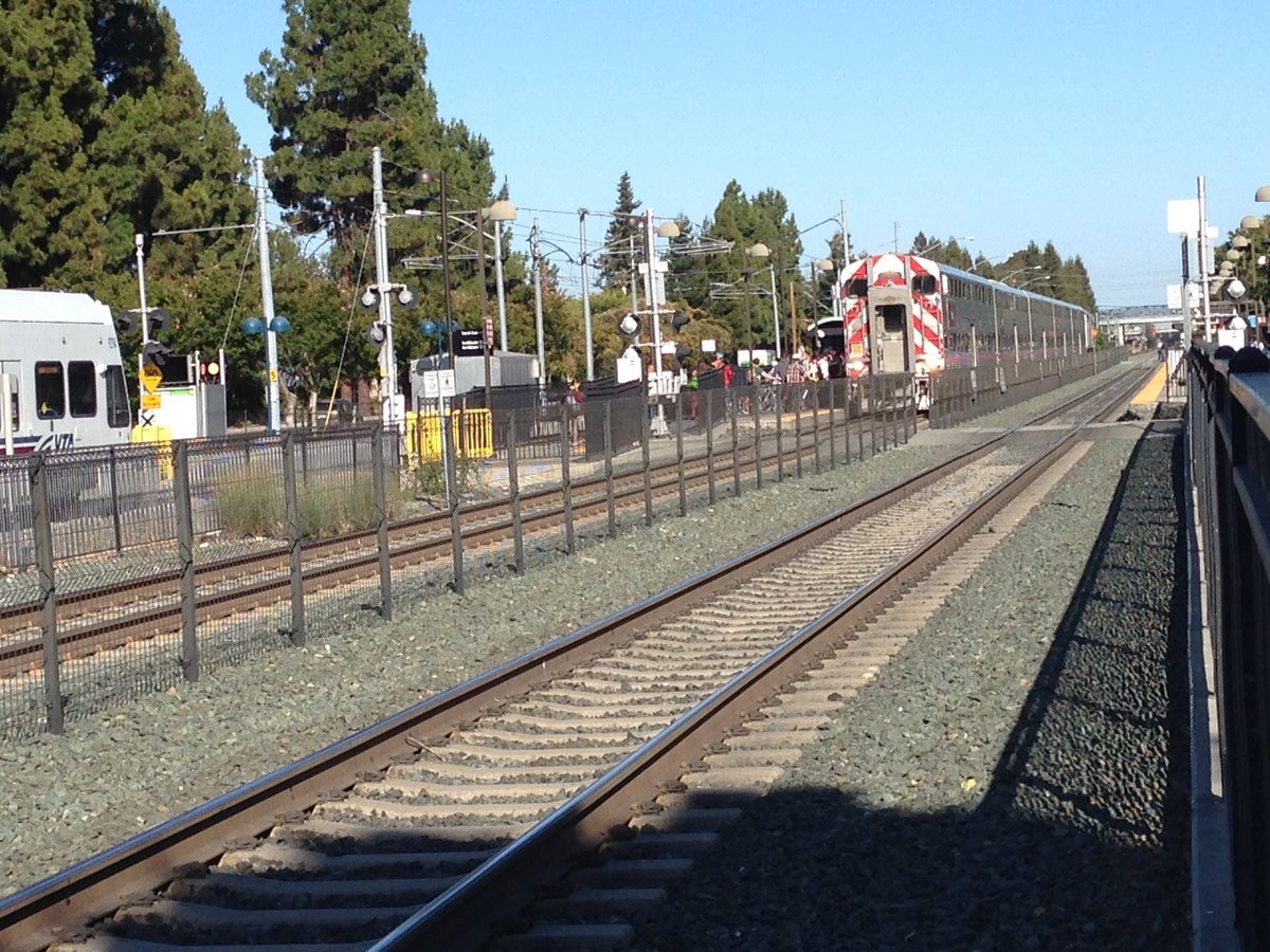 A Caltrain double-decker train stopped at the Mountain View station platform with passengers boarding on a sunny afternoon.