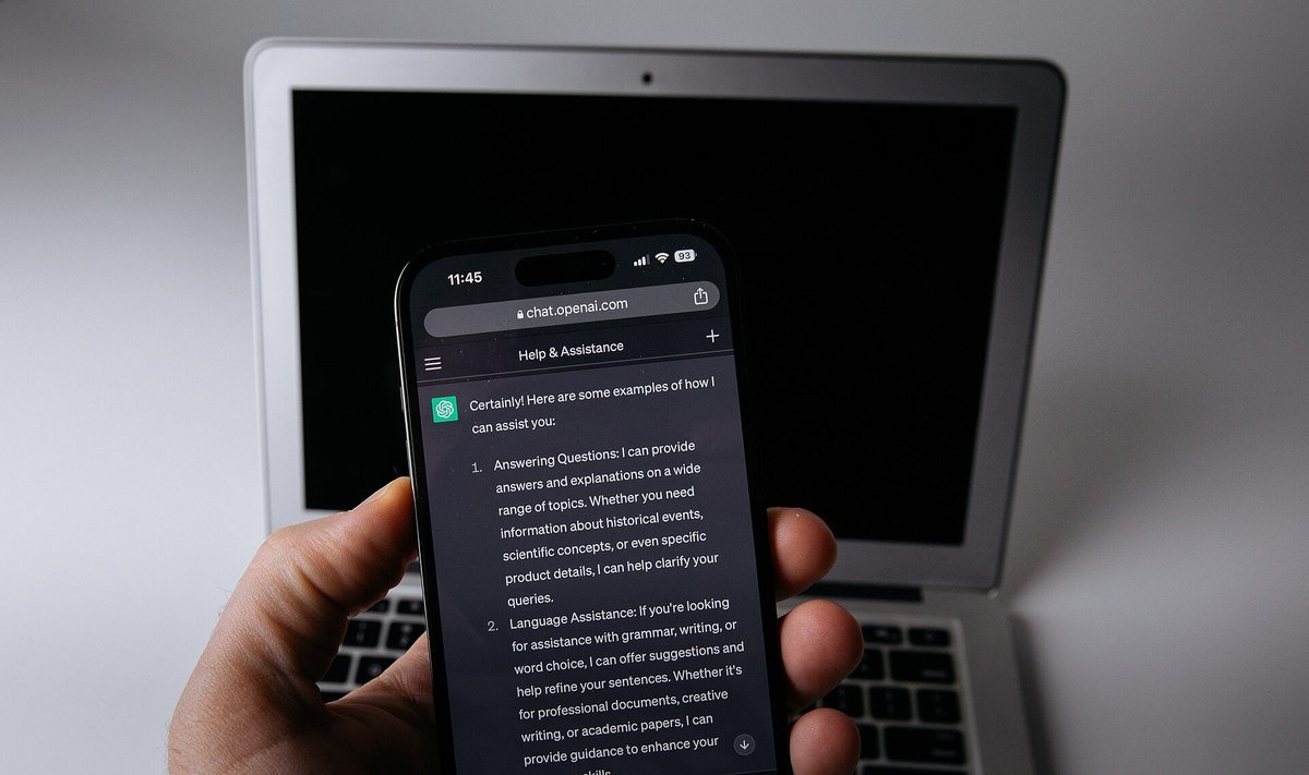 A hand holds a smartphone showing a ChatGPT conversation in dark mode in front of an open laptop