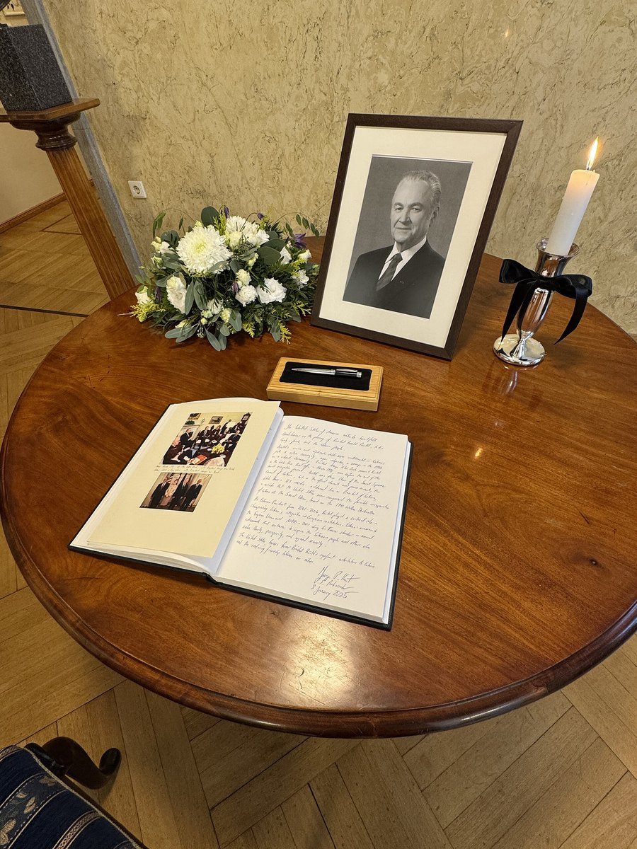 A condolence book lies open on a round wooden table beside a framed black and white portrait, a white flower arrangement and a candle