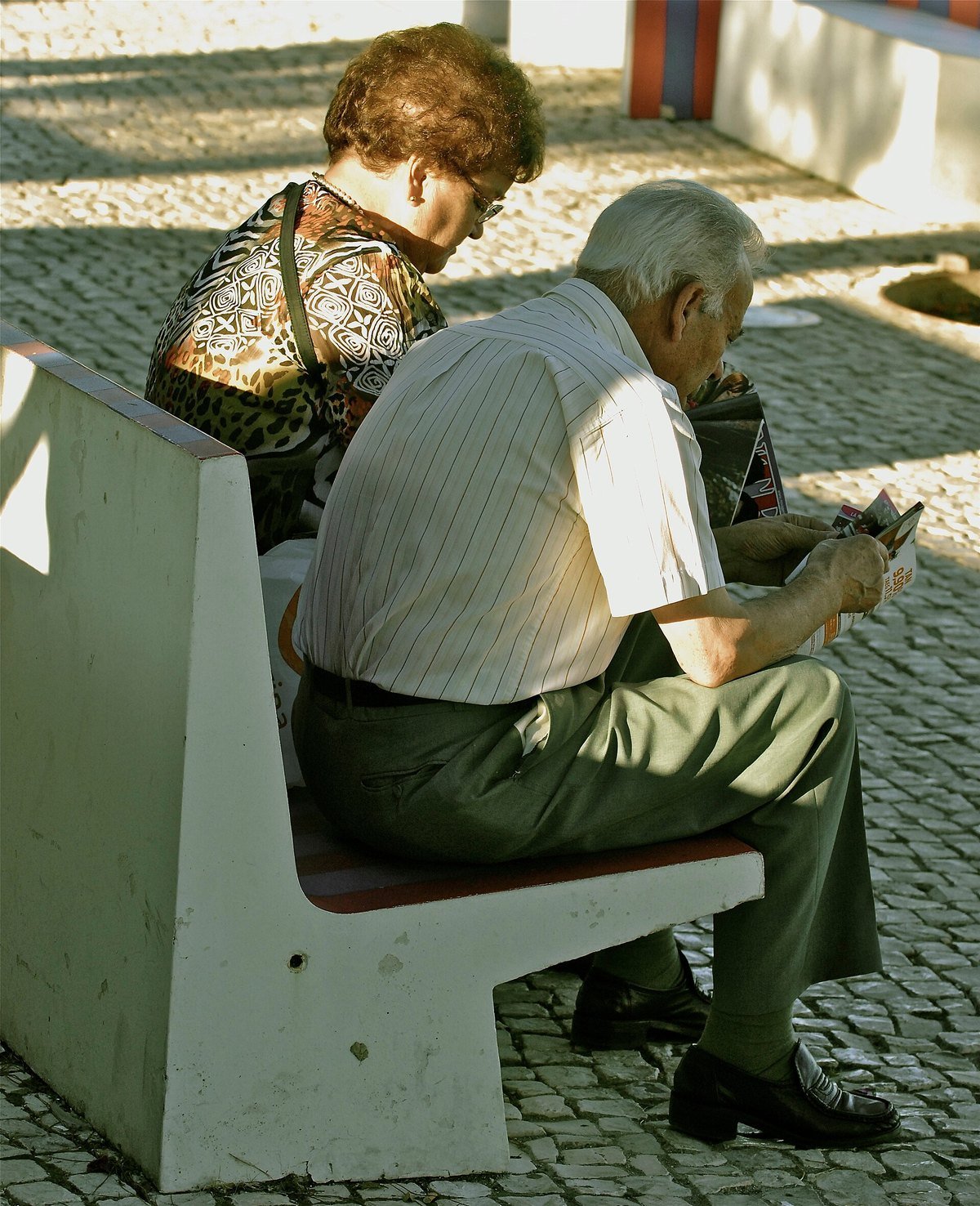An older man and woman sit side by side on a stone bench in the sun, each reading their own paper