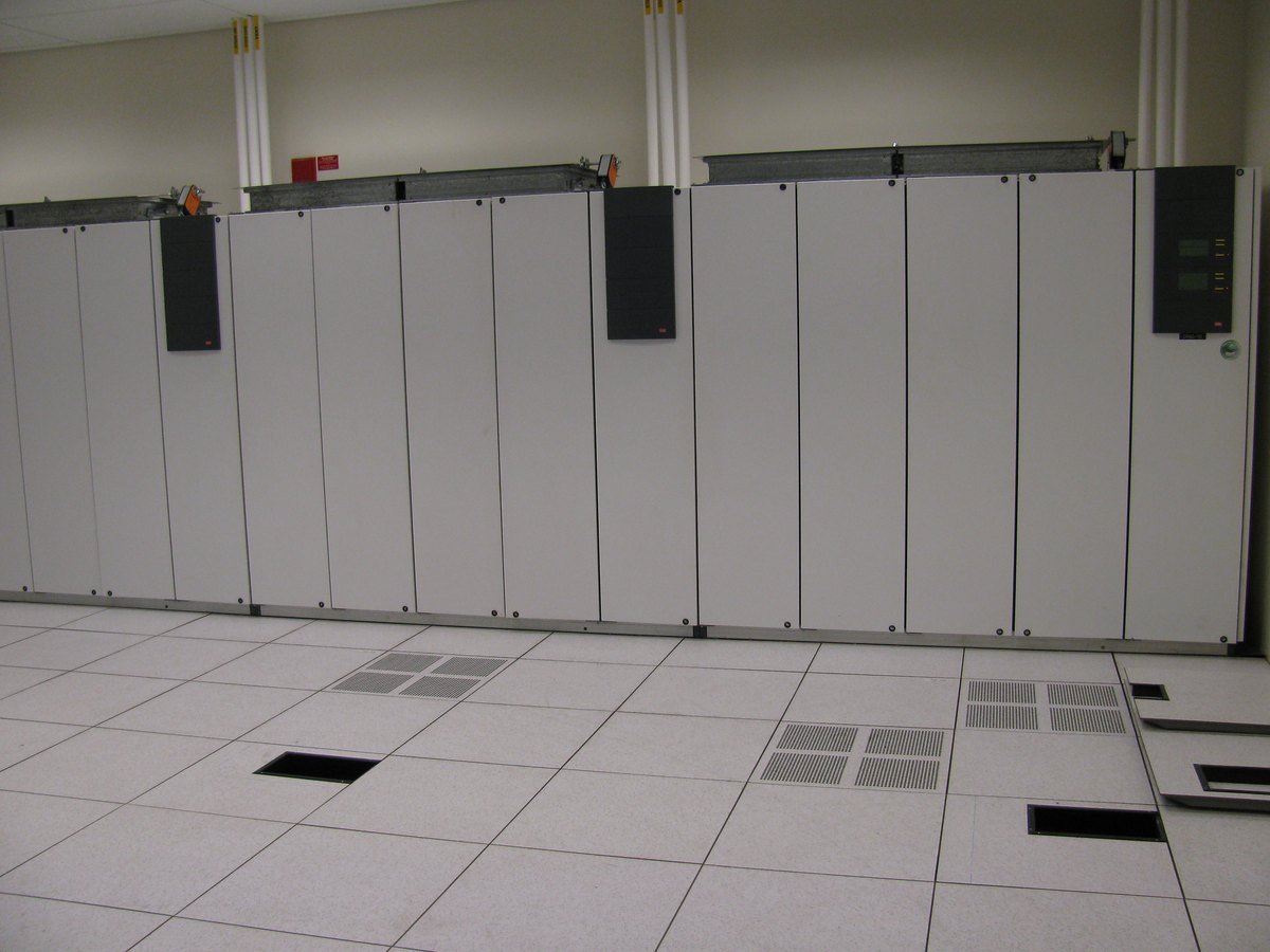 A row of large beige computer room air conditioning cabinets standing on a white raised floor with perforated vent tiles in a data center.