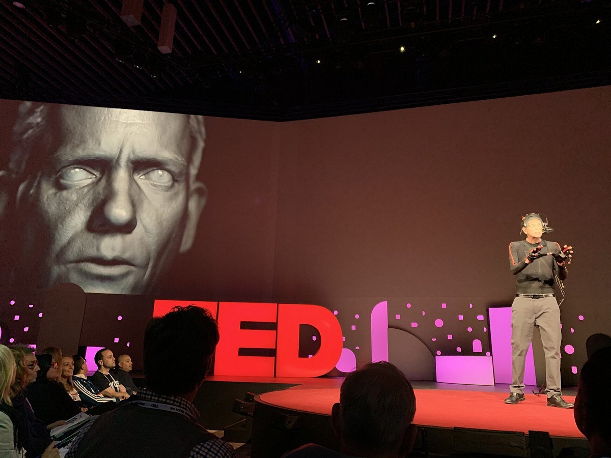 A large computer-generated face on a screen behind a speaker on the TED stage, with the audience in front