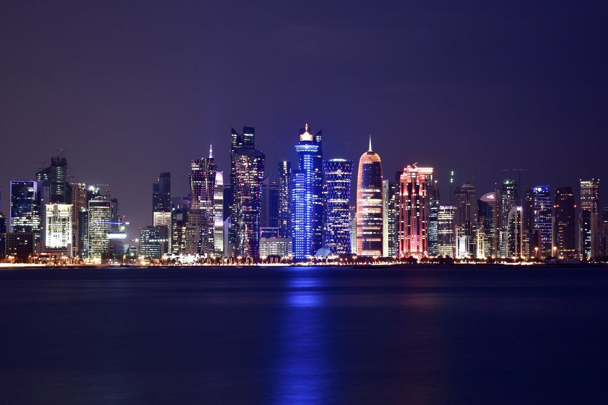 The West Bay skyline of Doha, Qatar at night, with brightly lit towers reflected in the water of the bay.