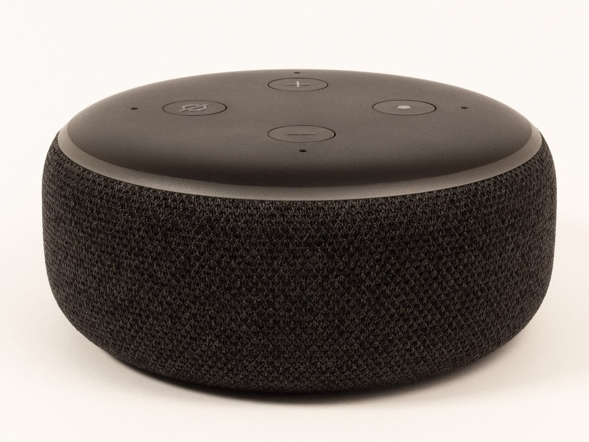A round dark gray Amazon Echo Dot smart speaker with four buttons on top, photographed on a white surface