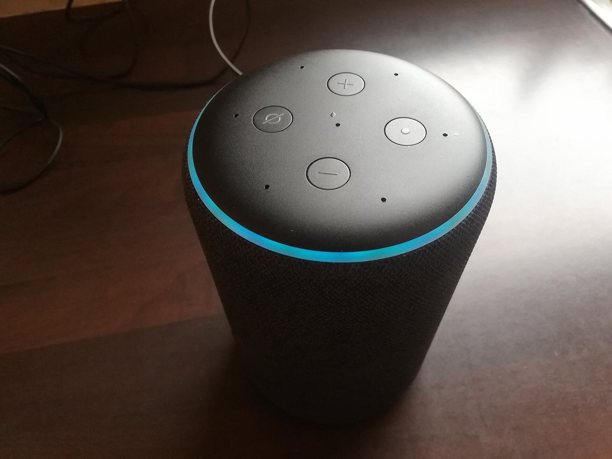 A dark gray Amazon Echo smart speaker on a wooden desk with a blue light ring glowing around its top edge