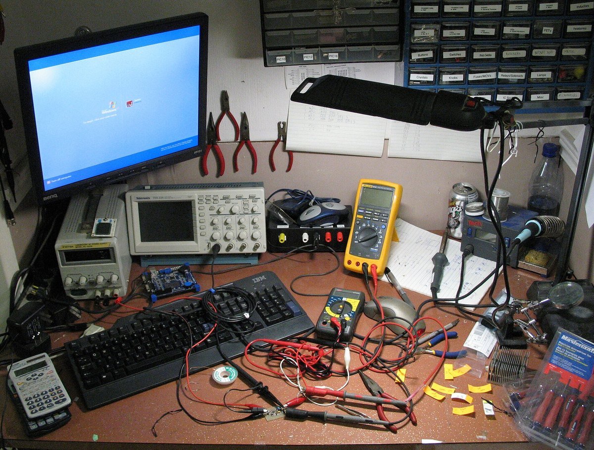 A crowded electronics workbench with an oscilloscope, two multimeters, a bench power supply, a soldering station, test leads and small parts drawers on the wall.