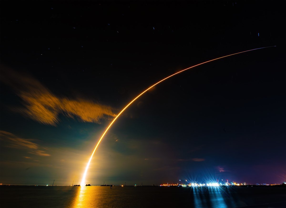Long-exposure photo of a SpaceX Falcon 9 rocket arcing across the night sky over water after launch from Cape Canaveral, carrying Starlink satellites.