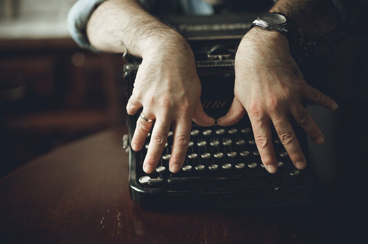 Close-up of a man's hands resting on the keys of an old black typewriter, with a wristwatch on one wrist