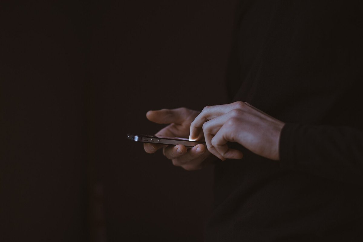 Two hands holding a smartphone in a dark room, a thumb scrolling the screen