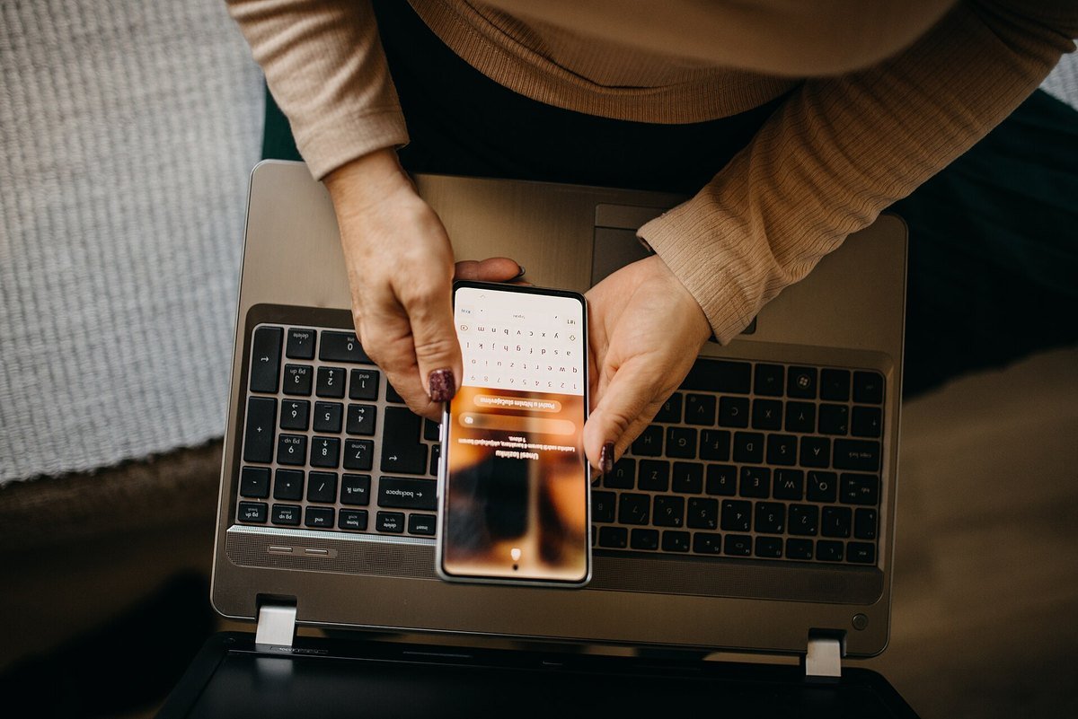 Seen from above, two hands hold a smartphone over an open laptop keyboard