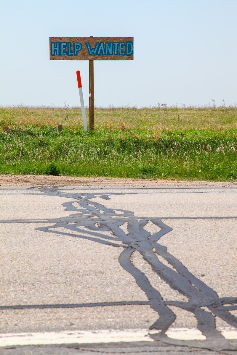 A hand-painted blue Help Wanted sign on a wooden post at the edge of a green field beside a patched rural road under a pale sky in Nebraska.