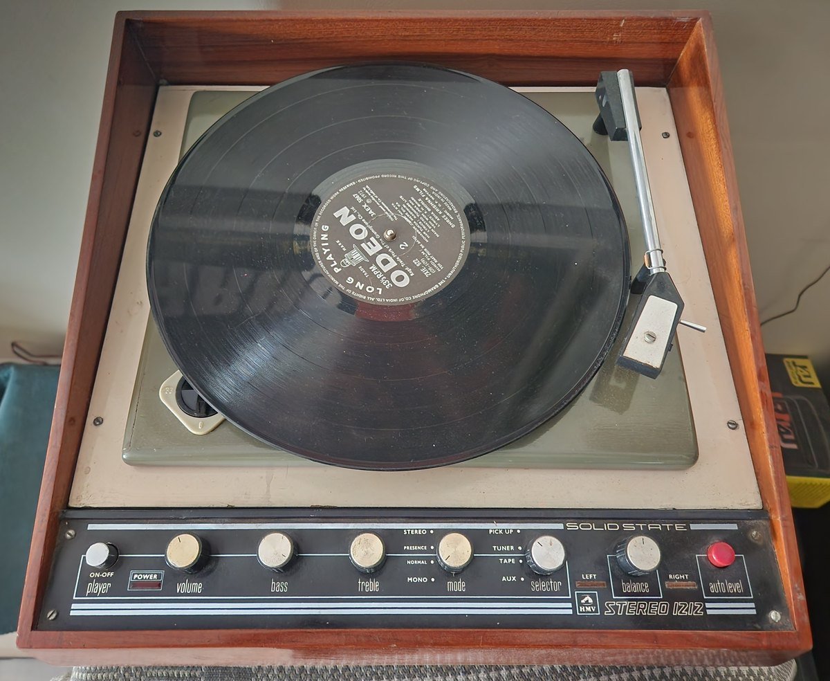 An HMV solid-state record player in a wooden case with a long-playing record on the turntable and control knobs for volume, bass and treble