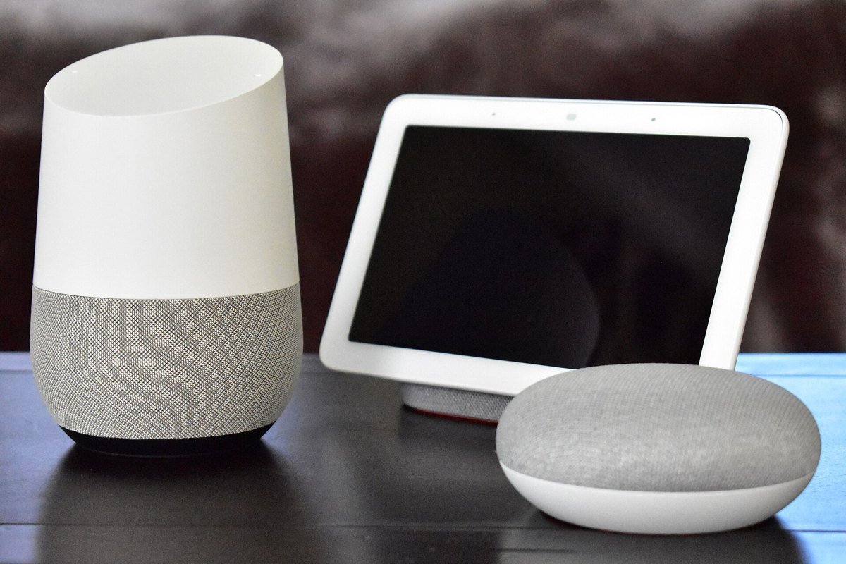 An original Google Home speaker, a Google Home Hub display and a Google Home Mini on a dark table