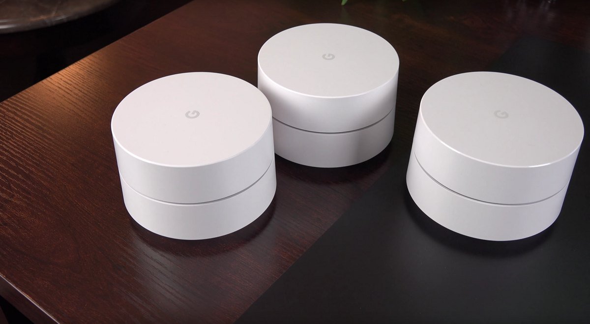 Three round white Google Wifi mesh router units sitting on a dark wooden table