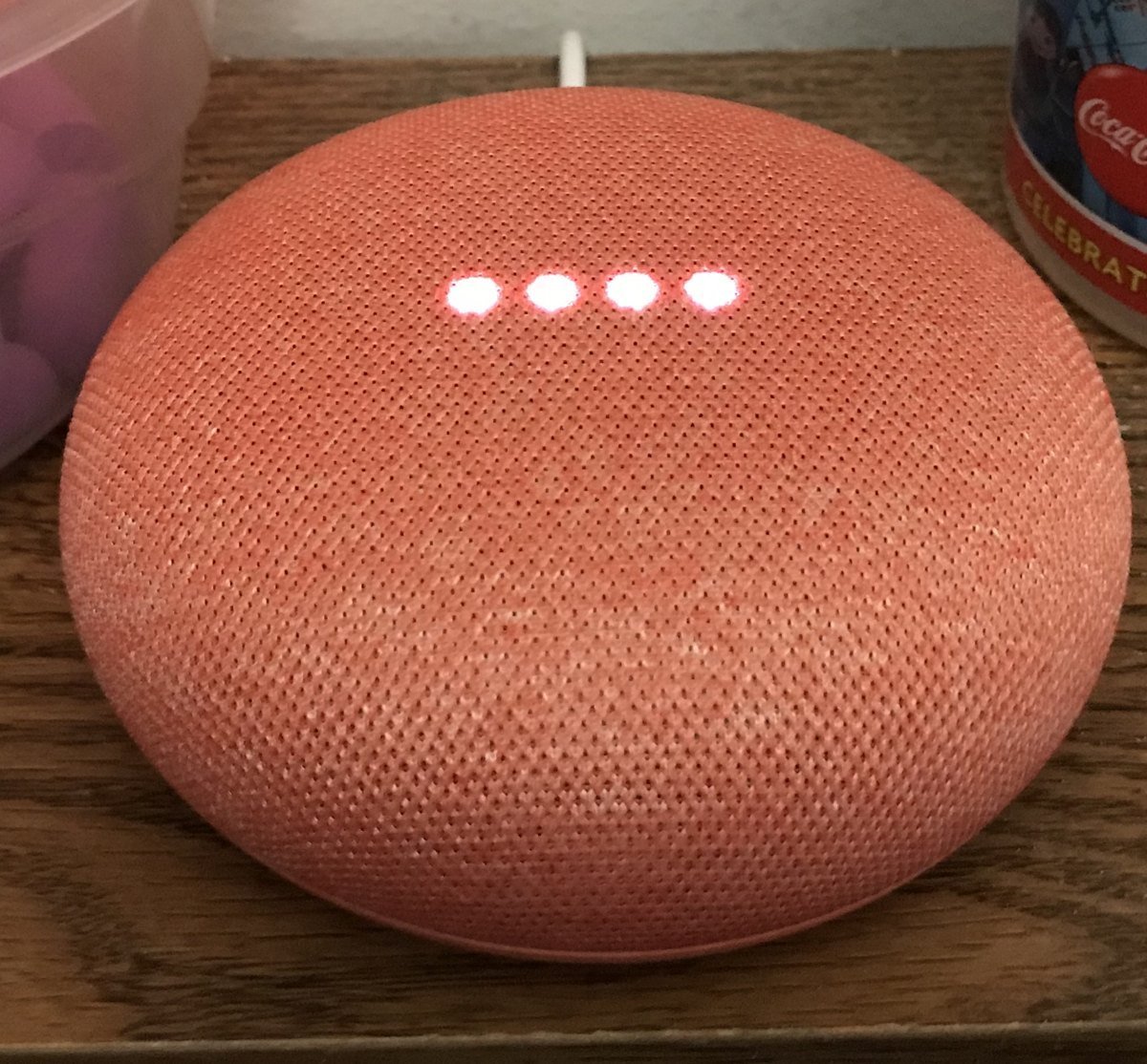 A coral Google Home Mini speaker on a wooden table with four small white lights glowing on top