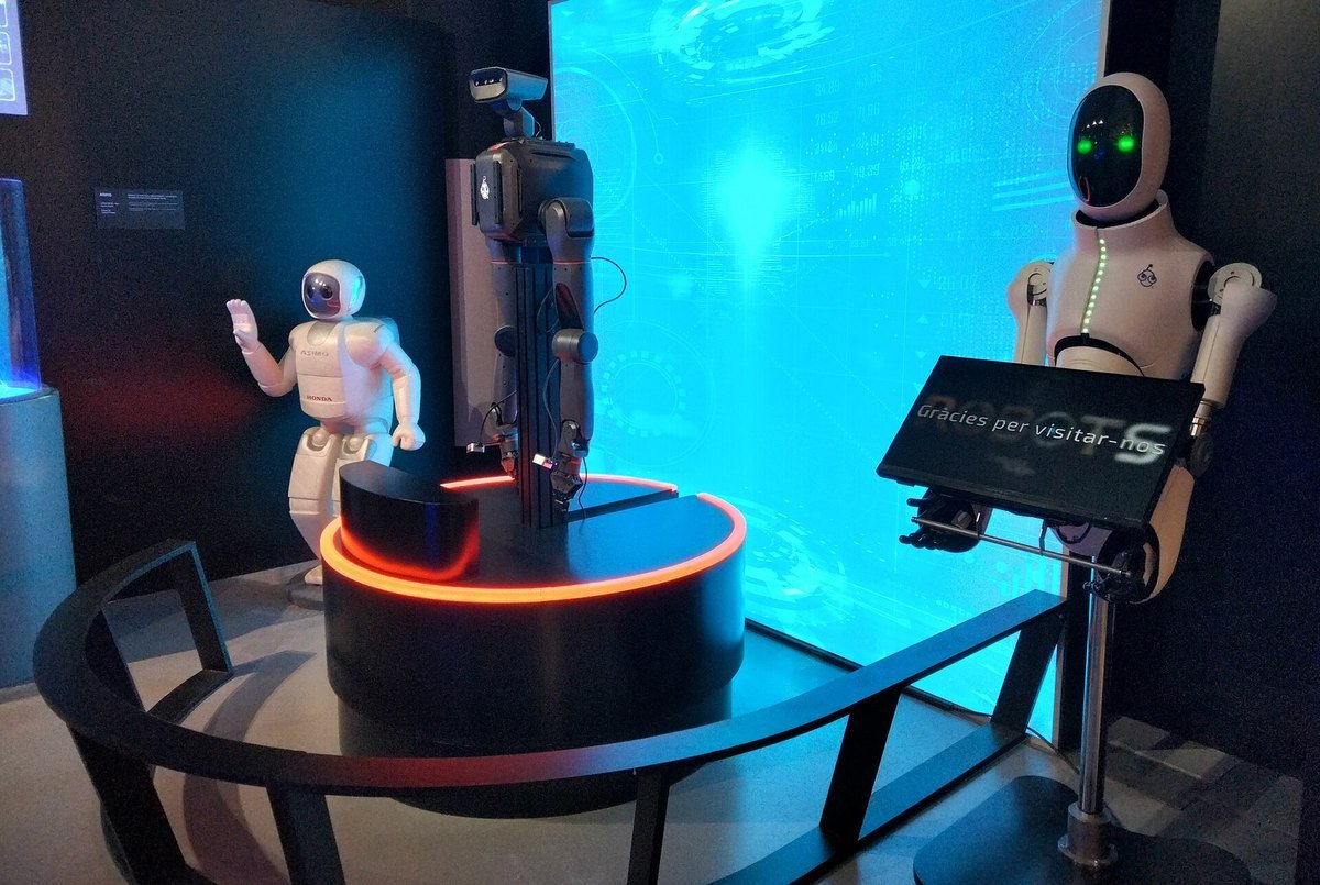 Three humanoid robots on display in a museum exhibition: a small white walking robot, a tall robot with arms on a lit pedestal, and a white humanoid with a dark face screen beside a sign.
