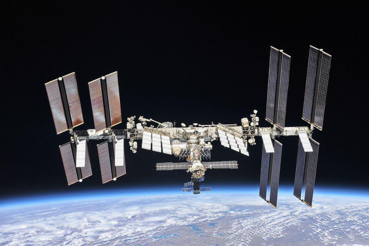 The International Space Station photographed from a departing Soyuz spacecraft, showing its long solar array wings and white radiator panels above the Earth.