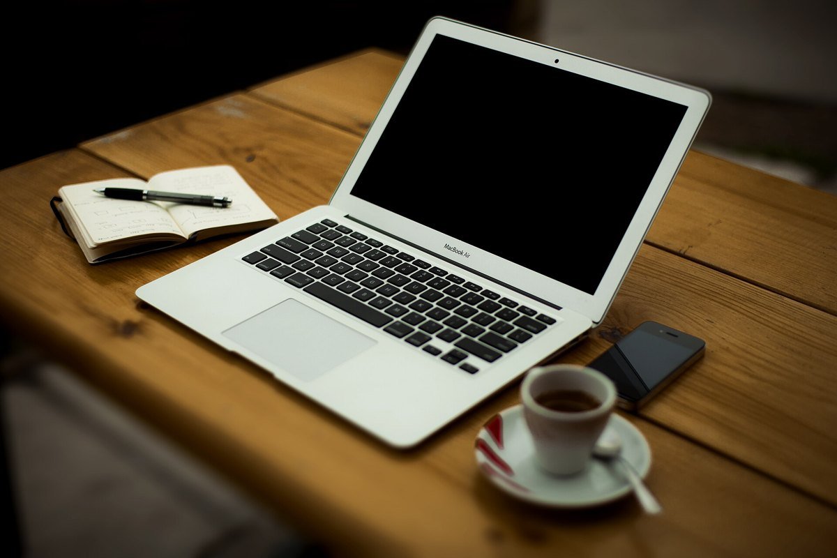 An open laptop on a wooden table at home, next to a notebook with a pen, a smartphone and a cup of espresso.