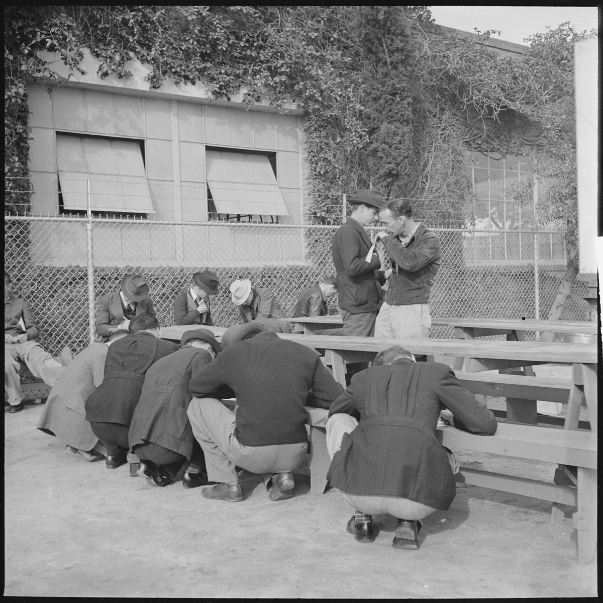 Black and white photograph of men in coats and hats crouching and leaning over outdoor wooden benches to fill out preliminary job applications beside a brick factory building in Los Angeles in 1940.