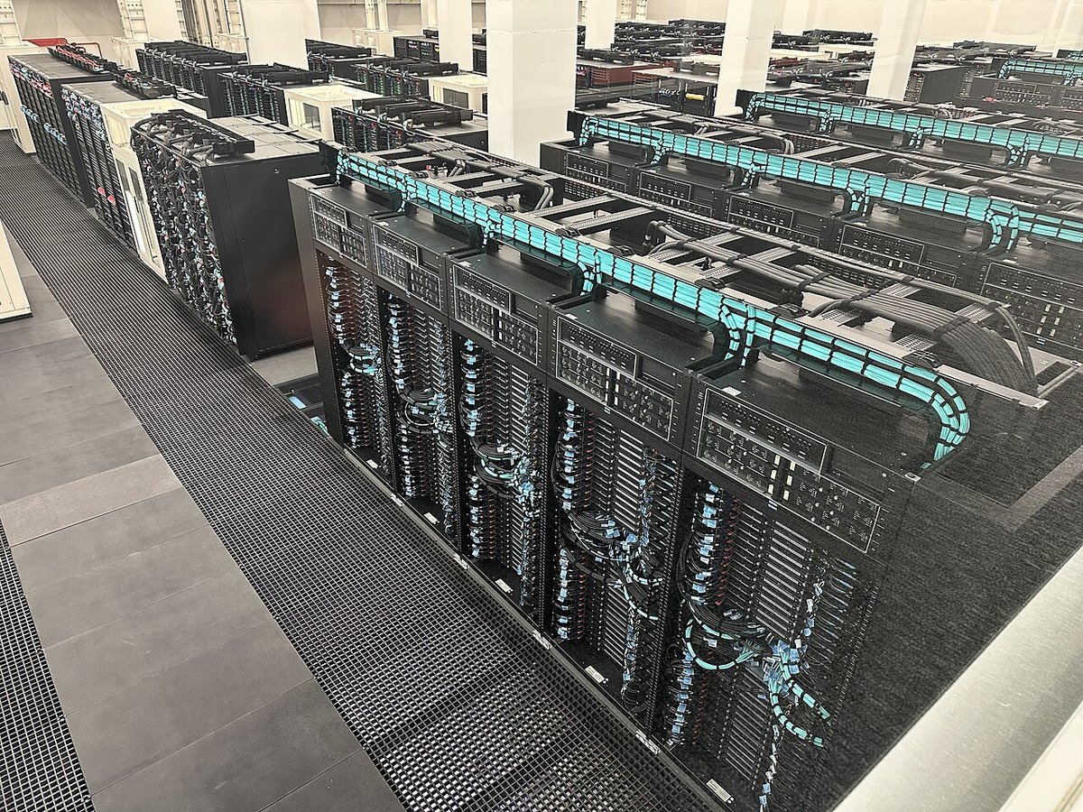 Long rows of black supercomputer server racks with teal cabling on a raised floor