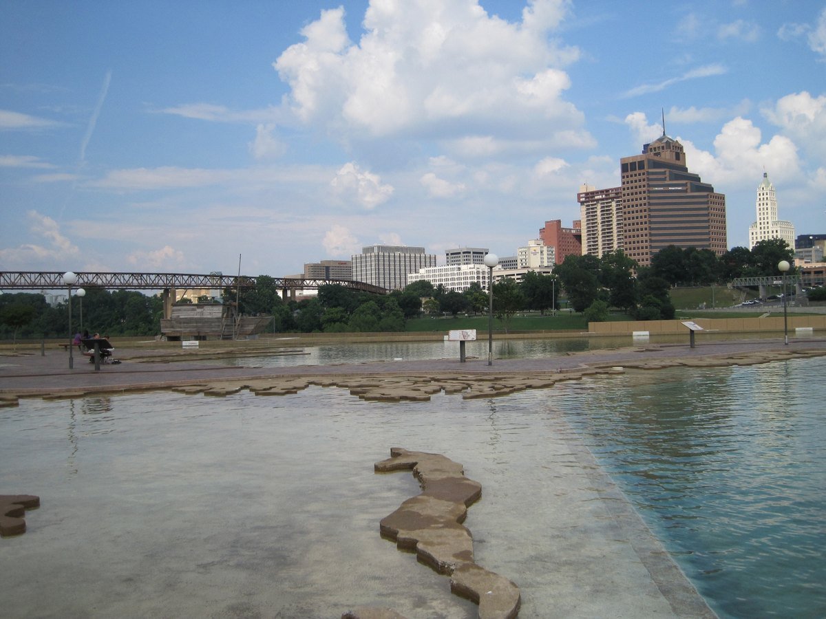 Downtown Memphis skyline seen across the shallow water model of the Mississippi River at Mud Island River Park, with a pedestrian bridge on the left and white clouds above.