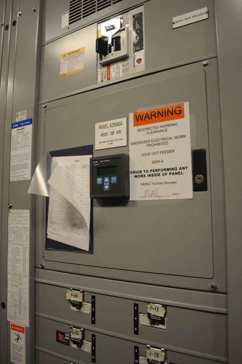 Gray electrical power panels in a computing center with an orange warning label about restricted working clearance and lockout steps before work inside the panel.