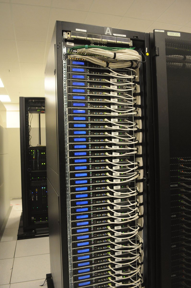 The back of a tall server rack in a data center, with neatly bundled white network cables running to rows of blue-lit servers.