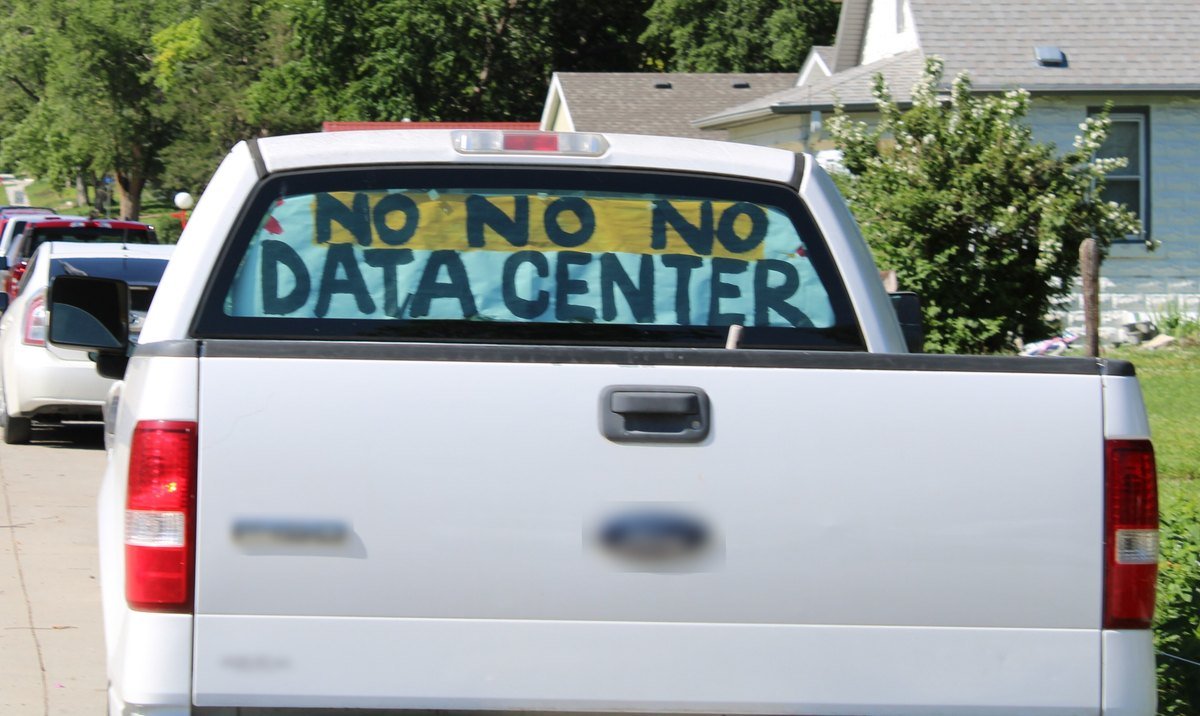 A white pickup truck parked on a residential street with a hand-painted sign in its rear window reading No No No Data Center.