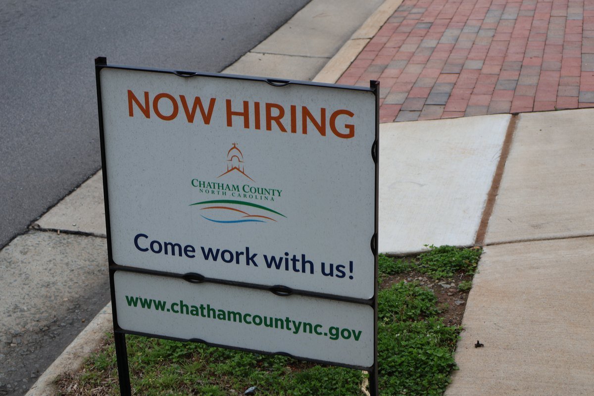 A white Now Hiring yard sign from Chatham County, North Carolina, reading Come work with us and the county website, staked in grass beside a brick sidewalk.